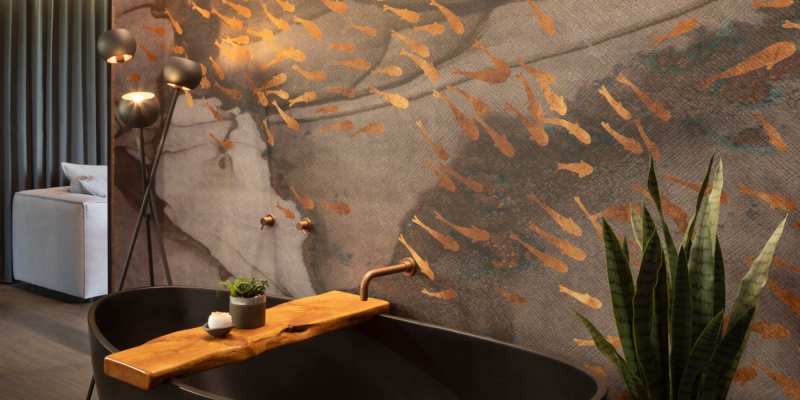 A black bathtub with a wooden board and copper faucet, accompanied by a potted plant and soap, set against a unique wall mural.